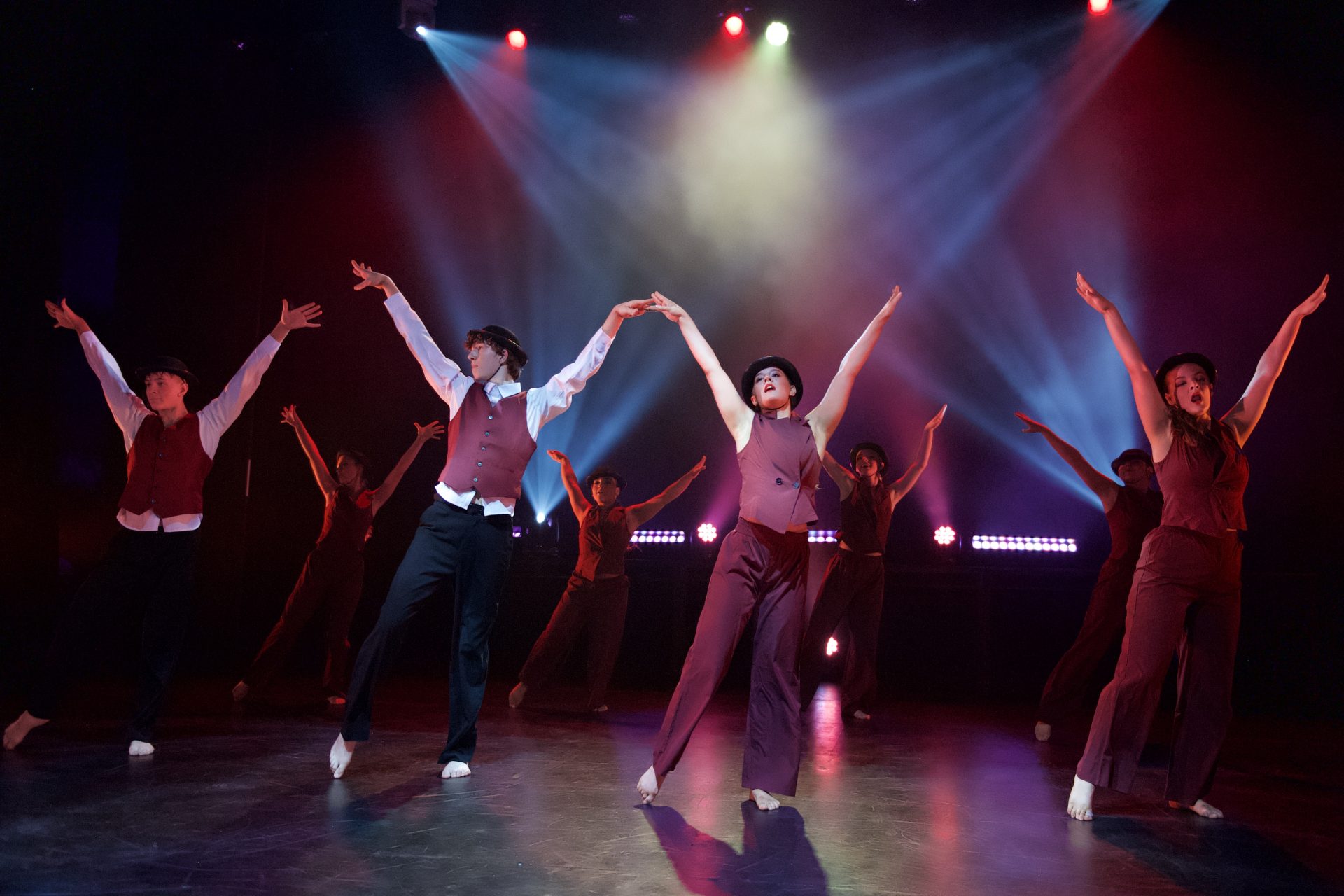 A group of dancers performing a synchronized stage dance under colorful theatrical lights, wearing matching vests and hats with arms raised in a dramatic pose.